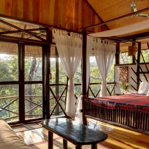 The image shows a cozy, rustic bedroom with a canopy bed, wooden furniture, large windows, white curtains, and a forest view outside.