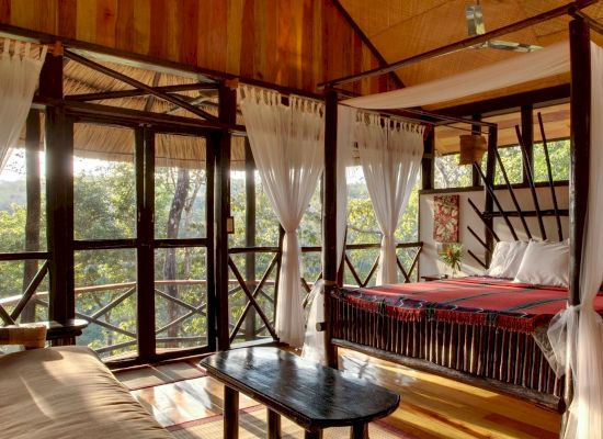 The image shows a cozy, rustic bedroom with a canopy bed, wooden furniture, large windows, white curtains, and a forest view outside.