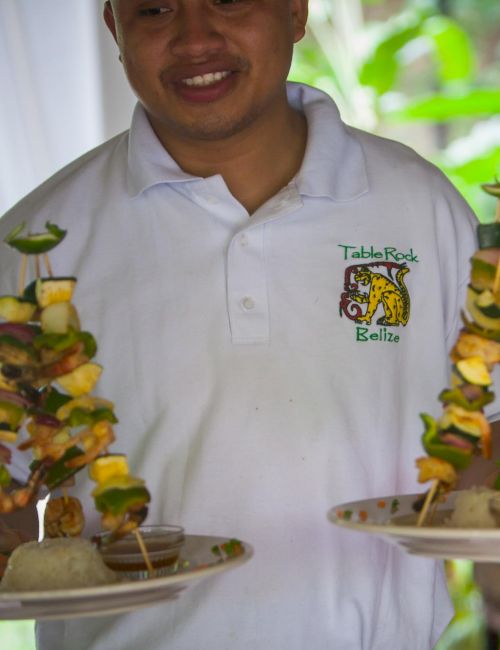 A person is holding two plates with creatively stacked skewers of food. They are wearing a white shirt with a logo.