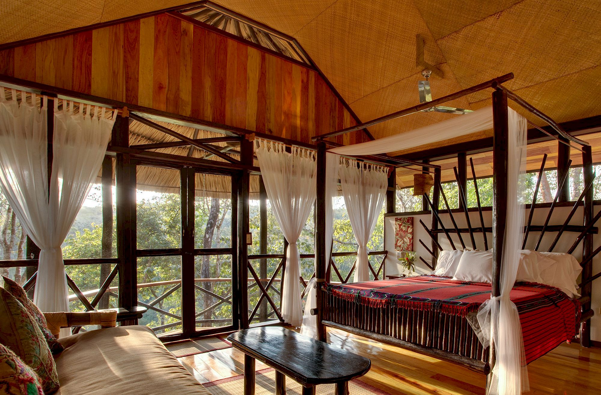 The image shows a cozy rustic bedroom with a four-poster bed, wooden walls, and large windows offering a scenic forest view.