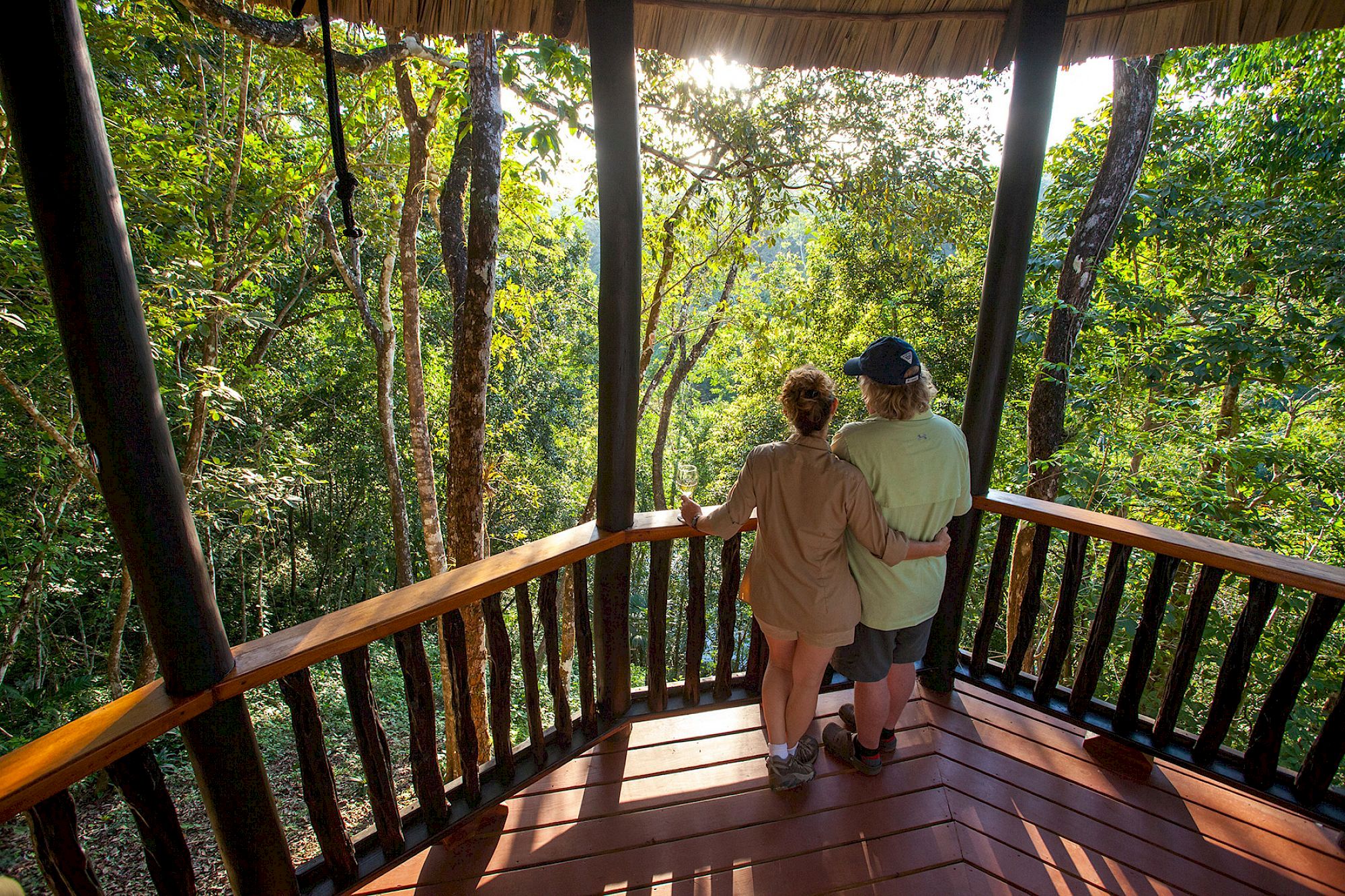 Two people stand on a wooden balcony, overlooking a lush, green forest under sunlight, creating a serene and peaceful scene.